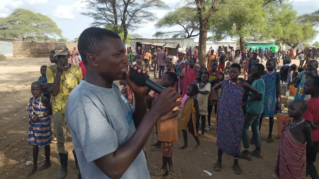 A health worker speaking into a microphone during a community awareness campaign, with residents gathered nearby.