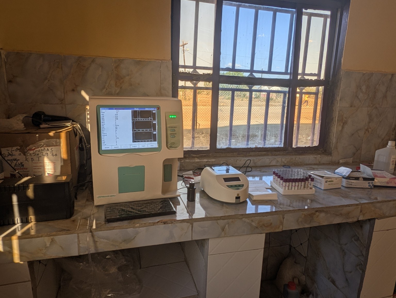 Diagnostic equipment and supplies on the tiled laboratory workbench at Nyakica Medical Centre.